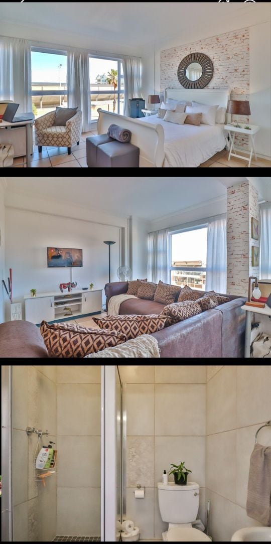 A bright and airy studio apartment features large windows allowing natural light to fill the space. A spacious bed is complemented by decorative cushions and a round mirror that adds a sense of depth. Comfortable seating is arranged to take advantage of the sunny view.