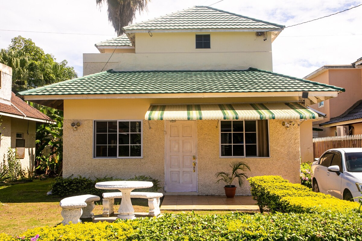 The house features a white exterior with a green-tiled roof and a welcoming front porch. A round table and stone benches are situated in the well-maintained garden, bordered by neatly trimmed hedges. Large windows allow natural light into the living spaces.
