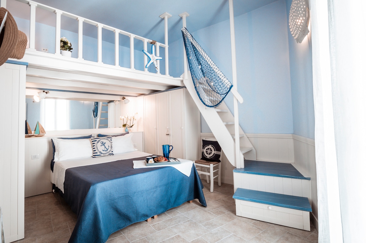 A bright bedroom features a spacious bed dressed in calming blue tones. An upper loft area is accessed via a white staircase, and a fishing net hangs decoratively from the wall. Natural light filters through a large window, illuminating the room's coastal-inspired decor.