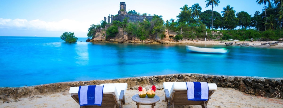 Two lounge chairs are positioned on a sandy beach, adorned with towels. The serene blue waters of the ocean gently lap at the shore, while a historic stone structure is visible in the background amidst lush greenery and palm trees.