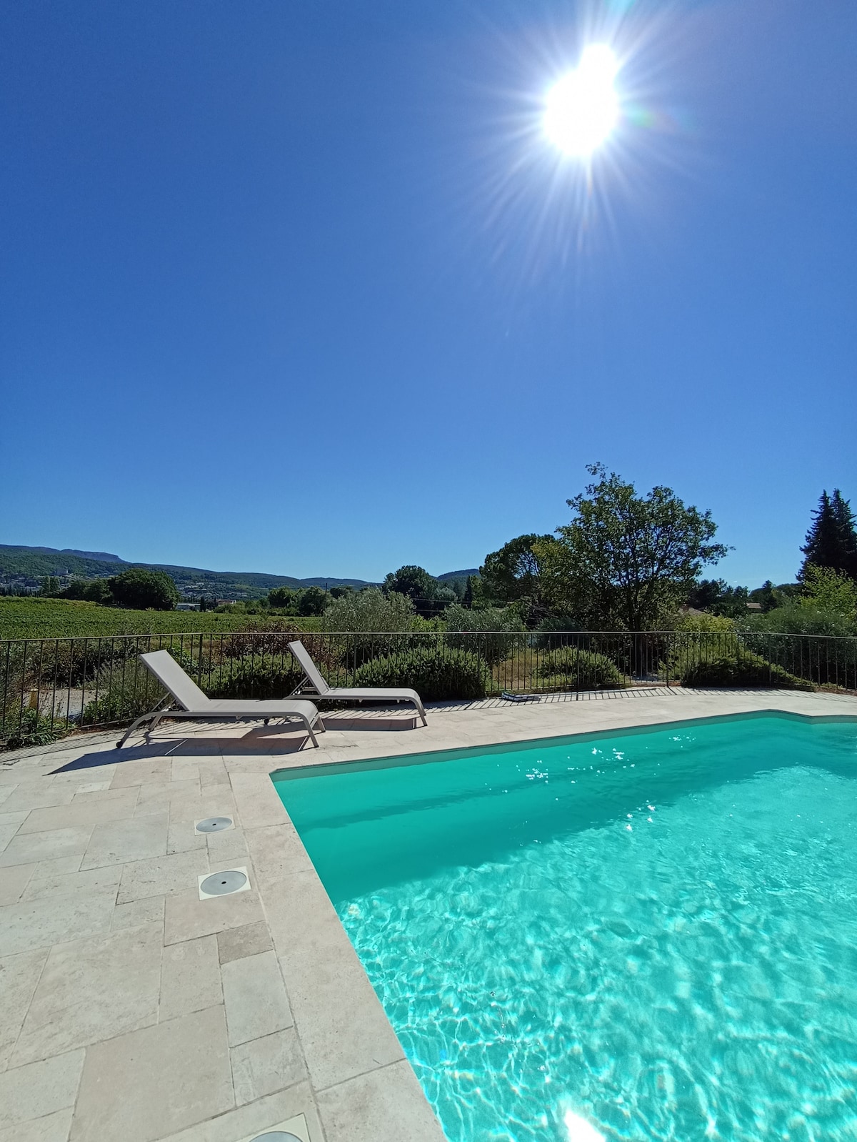 A private pool with clear blue water is surrounded by a stone patio and lush greenery. Two sun loungers are positioned nearby, offering a serene view of the distant hills under a bright sunlit sky.