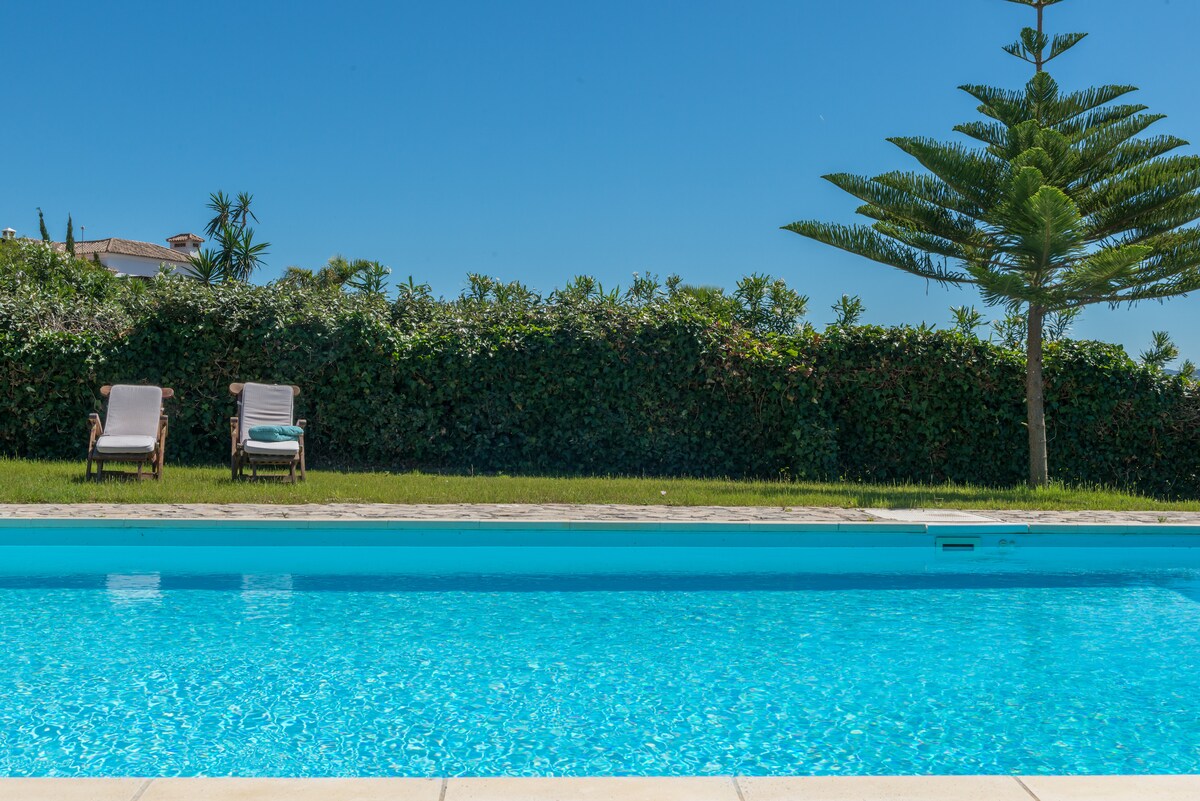 A serene private pool is surrounded by a well-maintained garden and hedges. Two sun loungers are positioned on the grassy area beside the pool, providing an inviting space for relaxation under a clear blue sky.