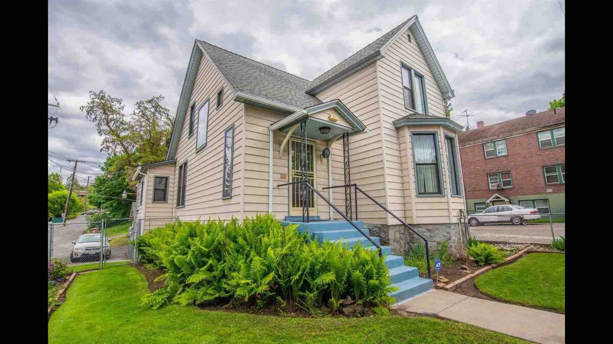 A charming Victorian home with a light-colored exterior features distinctive architectural details. The entrance is marked by a set of blue steps leading to the front door. Lush greenery surrounds the property, enhancing its suburban appeal.