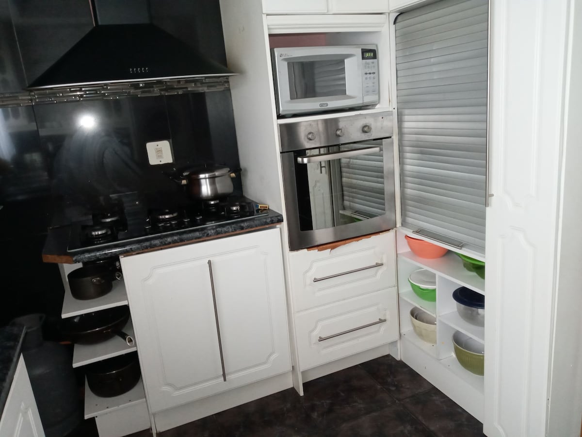The kitchen features modern appliances including a stove, microwave, and oven within white cabinetry. Open shelves display an array of colorful bowls. The countertop is dark, complementing the overall design, while the space is illuminated, offering a functional cooking environment.