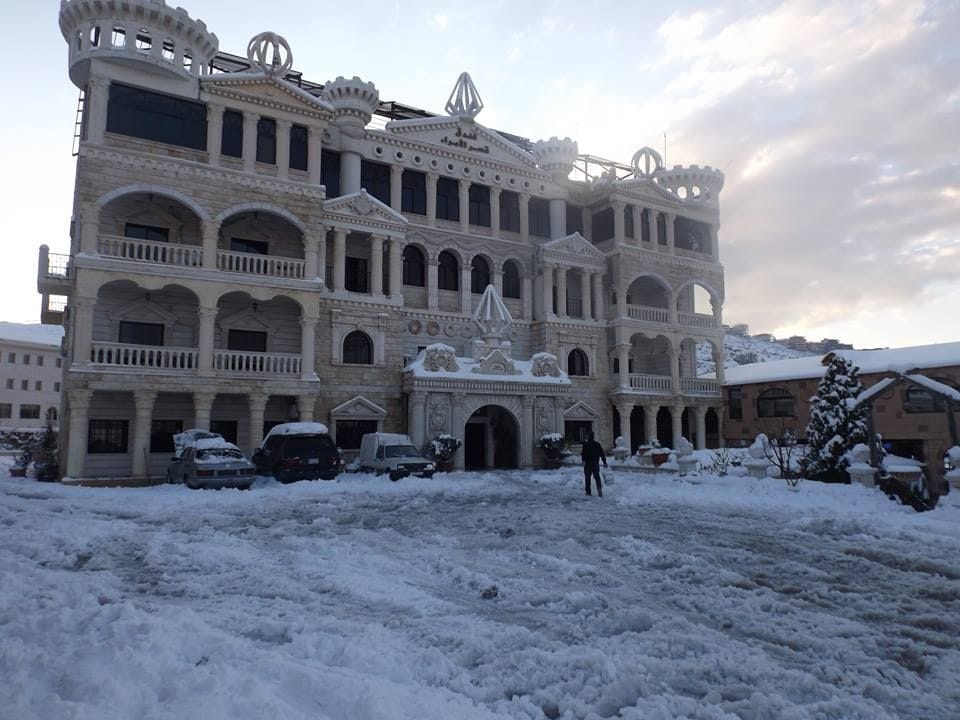 A grand stone building with ornate architectural details is surrounded by a blanket of snow. The entrance features large arches and decorative elements. Vehicles are parked in the snow-covered lot, and a person is visible walking on the snowy ground.