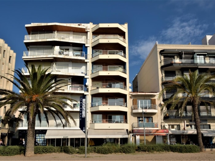 The building features a modern architectural design with multiple balconies and large windows. Palm trees are visible in front, complementing the coastal setting. Street-level establishments offer convenience. The structure is part of a vibrant area, with a clear blue sky in the background.