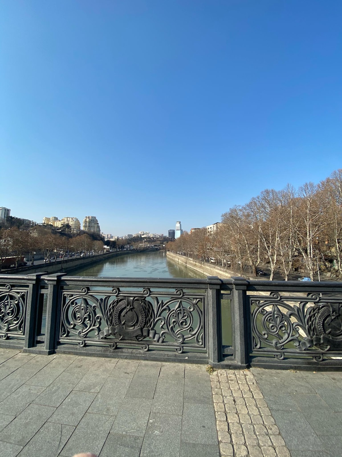 The image captures a view of the Kura River with clear blue skies above. The ornate railing of a bridge is visible in the foreground, complementing the serene waters. Leafless trees line the riverbank, and urban buildings are seen in the distance, contributing to the cityscape.