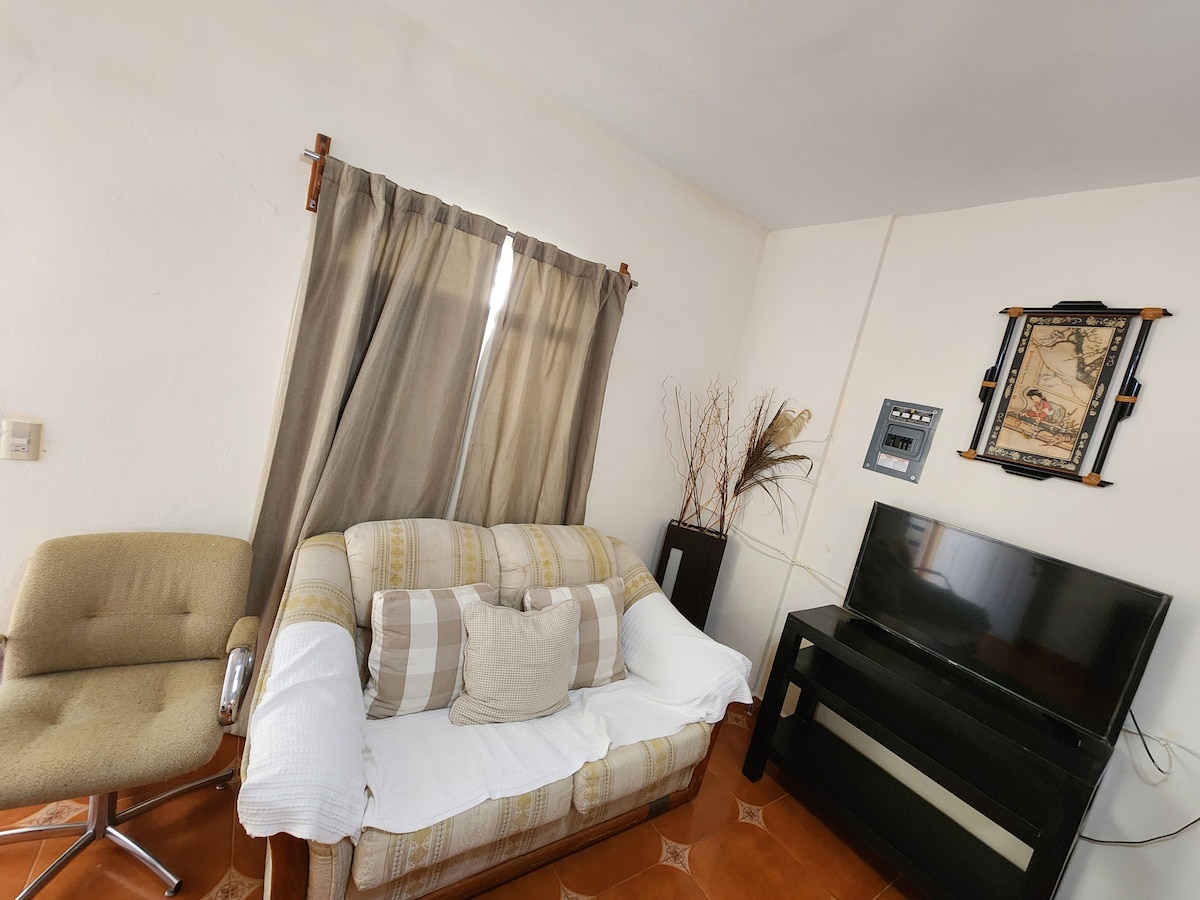 A living room area features a comfortable sofa with accent cushions and a plush white throw. A sleek black TV stand holds a flat-screen television. Natural light filters through light brown curtains, and a decorative piece is displayed on the wall, complemented by a chair in the corner.