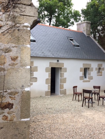 The exterior of the house is shown, featuring a white façade with stone accents and a gray slate roof. A gravel area is visible, with a small outdoor seating arrangement consisting of several wooden chairs and a table. Surrounding greenery enhances the natural setting.