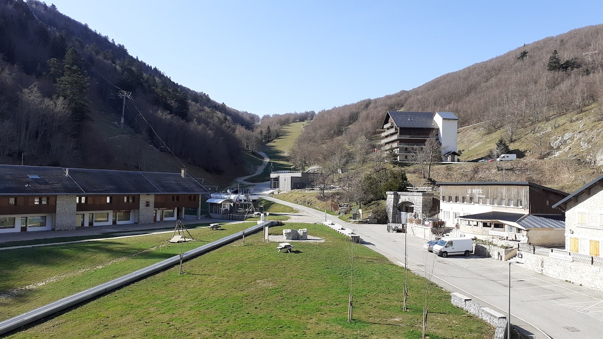 A broad view of the surrounding mountainous landscape, featuring a ski slope on the left and various buildings including accommodations. Green grass occupies the foreground, while tree-covered hills rise in the background under a clear blue sky.