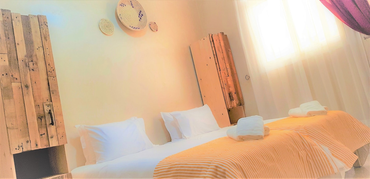 The room features two spacious beds dressed in crisp white linens, accompanied by folded towels. Wooden cabinets made from recycled materials stand beside the beds, with textured wall hangings adding to the warm decor. Natural light filters through sheer curtains, brightening the space.
