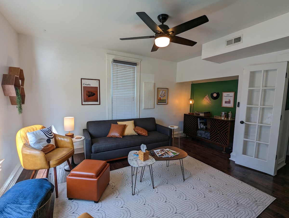 The living room features a modern gray sofa accompanied by orange and blue accent pillows. A round coffee table sits in the center, with a decorative item displayed atop it. A ceiling fan provides airflow, while bright light filters through a window, creating a welcoming space.