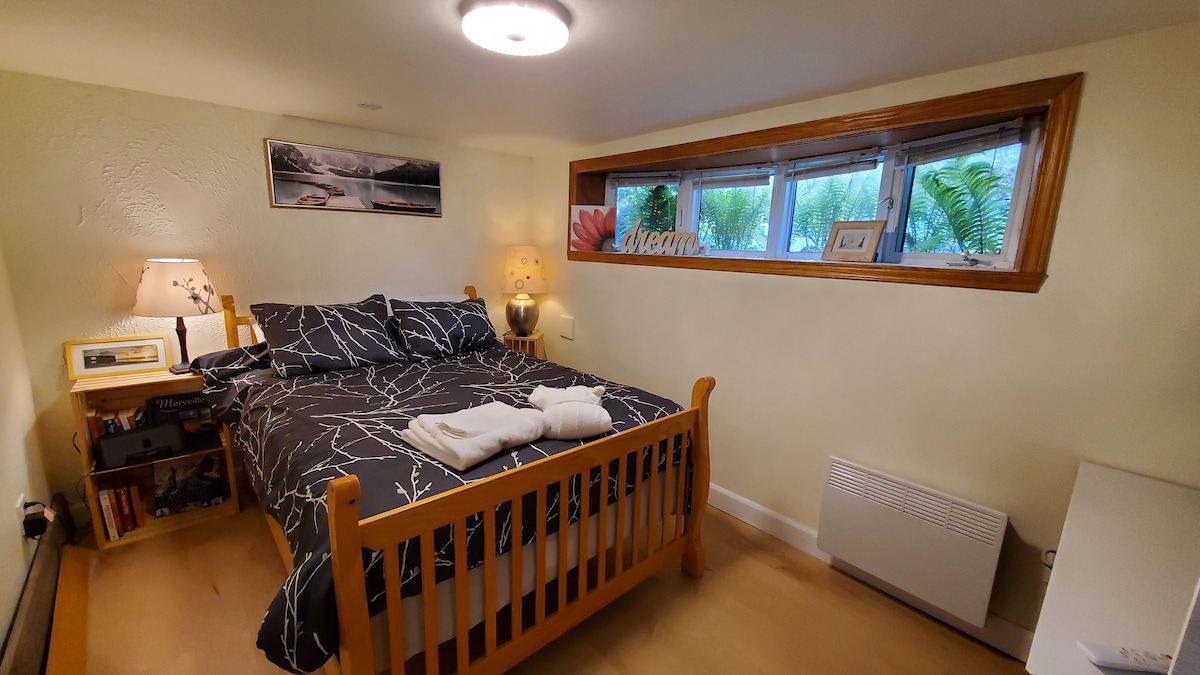 A well-appointed sleeping area features a wooden bed with a dark patterned duvet. Natural light filters through a wide window, complemented by soft lighting from a bedside lamp. A small nightstand and a white dresser are included, adding functional elements to the cozy space.