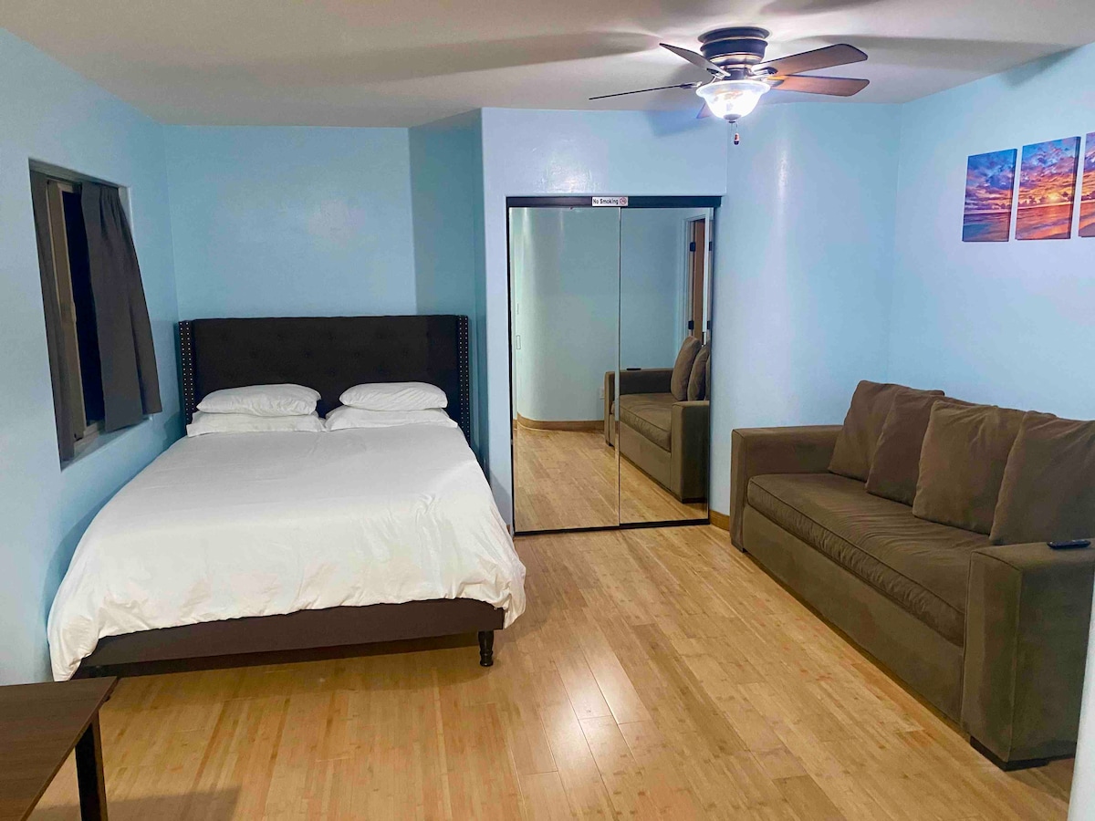 The studio apartment features a queen bed with white bedding, a west elm sofa bed in a neutral tone, and a small table. A mirrored closet reflects light and adds depth to the space. Soft blue walls create a calm atmosphere.