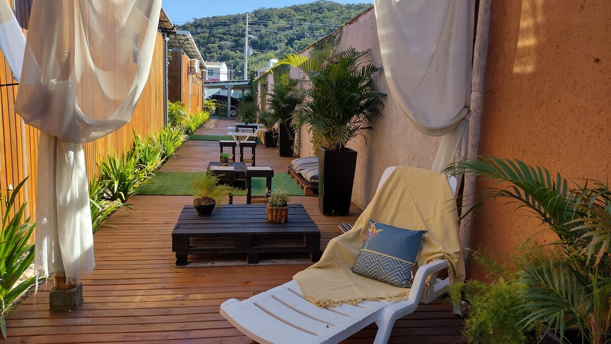A serene outdoor lounge area is highlighted by wooden decking, featuring sun loungers and low tables. Potted plants provide greenery along the pathway, which leads to a cozy atmosphere framed by soft drapes and natural surroundings.