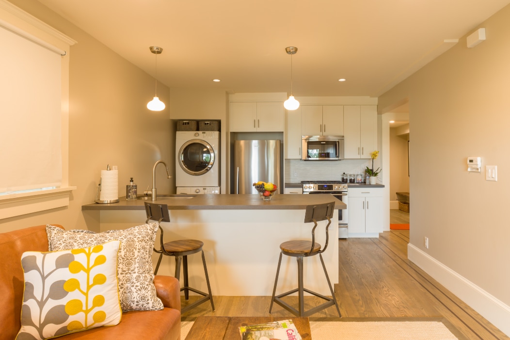 A modern kitchen and living area are showcased, featuring a central island with two stools. Stainless steel appliances and a washer-dryer unit are visible. Light-colored cabinetry and a cozy sofa with decorative pillows contribute to a welcoming environment. Natural light filters through a nearby window.
