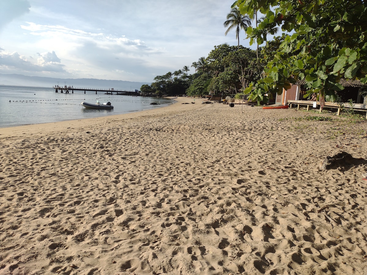 A sandy beach extends along the shoreline, with gentle waves lapping at the shore. Nearby, scattered trees provide shade, and a small boat is anchored close to the beach. In the distance, a wooden pier juts into the water, framed by lush greenery.