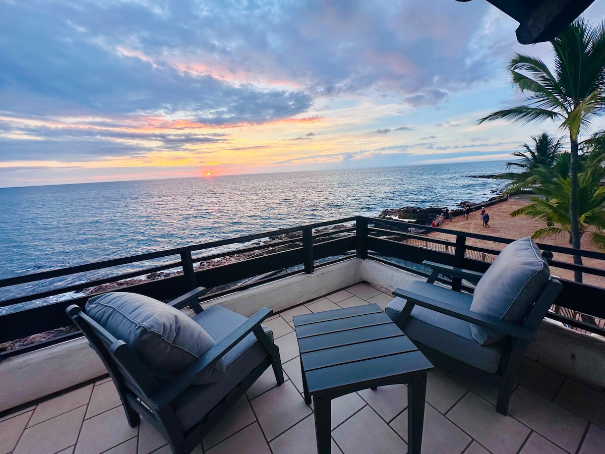A wrap-around lanai features two cushioned chairs and a small table, overlooking the ocean. The sunset casts warm colors across the sky, reflecting on the water below. Palm trees sway nearby, enhancing the serene coastal setting.