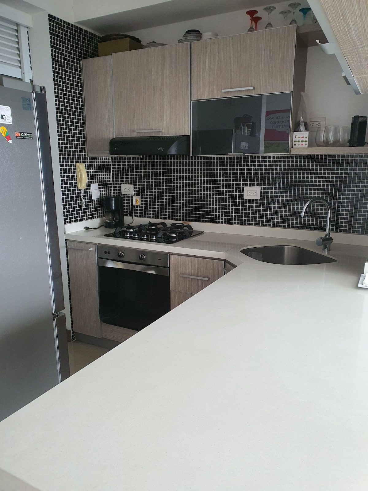 A compact kitchen area features light wood cabinetry and dark mosaic tiles. Stainless steel appliances, including a gas stove and refrigerator, are present. A modern sink is integrated into the spacious countertop, providing ample preparation space for cooking activities.