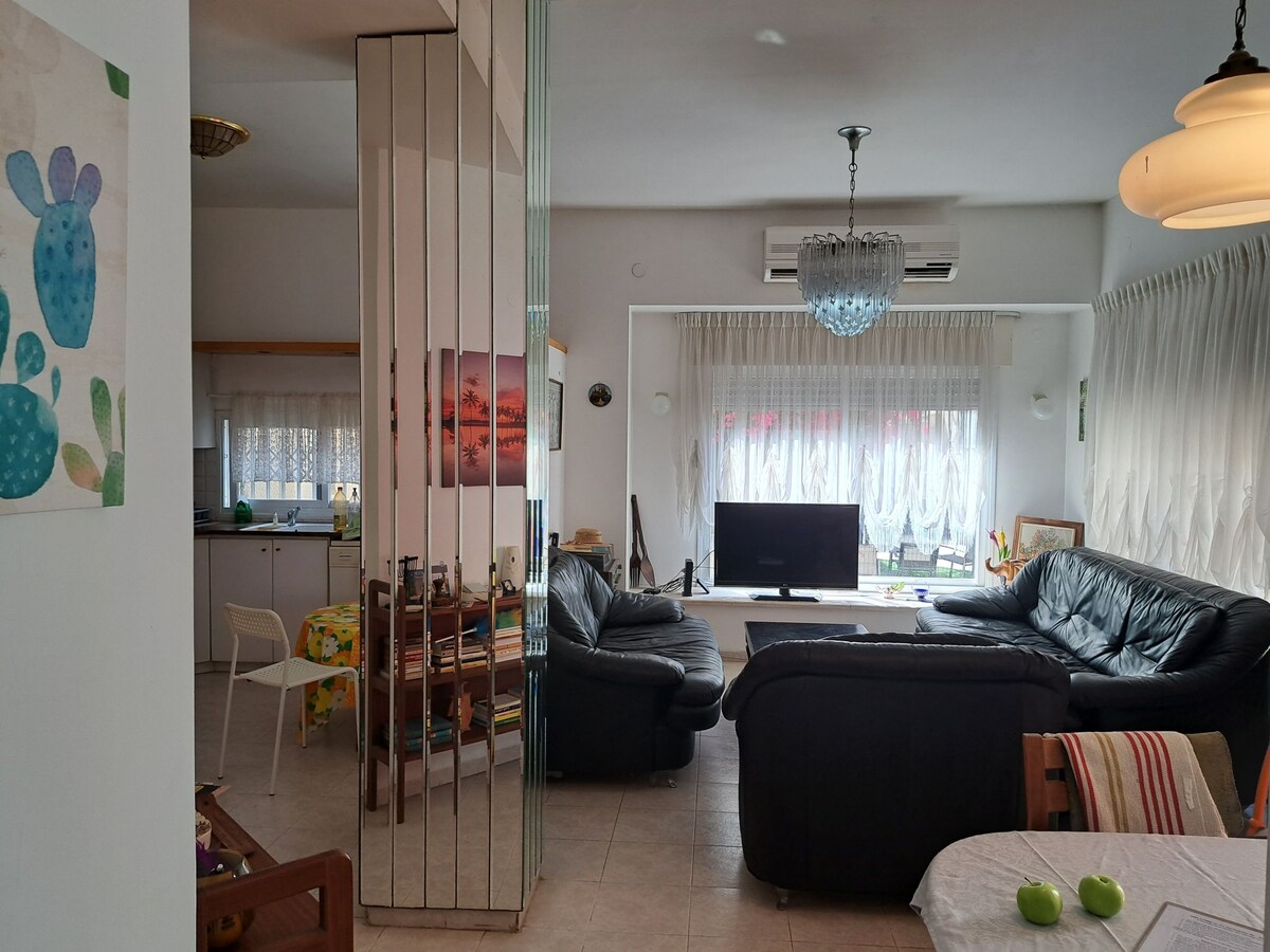 A spacious living area features two black leather sofas facing a flat-screen television. Light streams through sheer curtains, illuminating the room. A dining table with colorful tableware and artwork is visible, along with a mirrored wall that reflects the space.