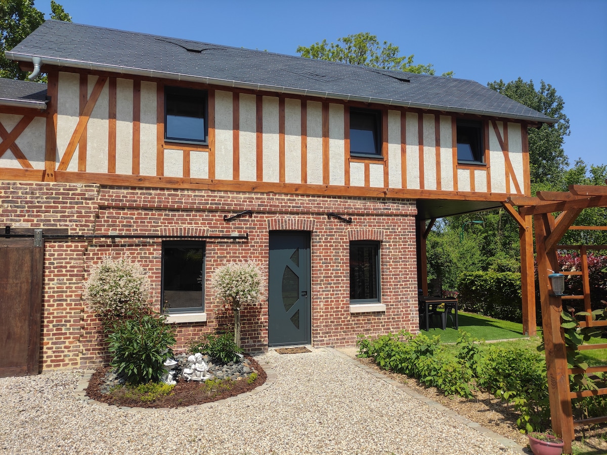 The exterior of the gîte features a charming blend of brick and wooden panels, framed by greenery. A gravel pathway leads to the entrance, flanked by ornamental shrubs. A covered porch area extends over the entrance, providing access to the surrounding garden space.