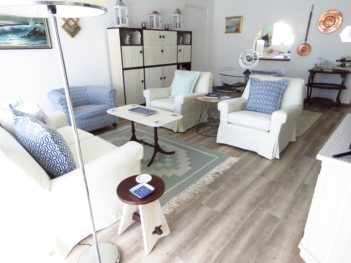 A light-filled living area features two white armchairs and a blue-patterned couch, arranged around a small wooden table. A patterned rug underfoot adds warmth, while a standing lamp provides additional light. Decorative pieces adorn the shelves, creating a comfortable and stylish ambiance.