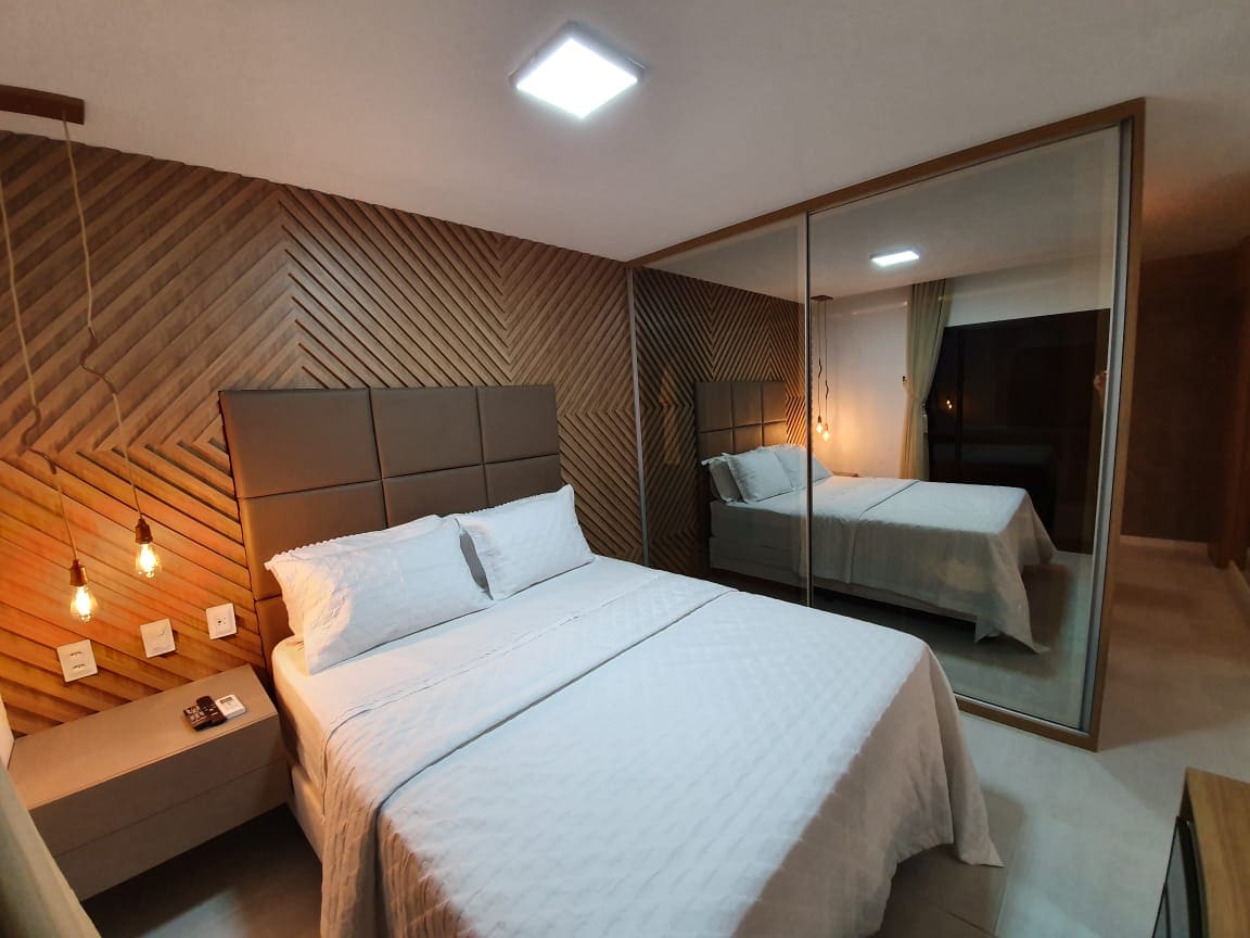 A spacious sleeping area features a large bed with a light-colored duvet, complemented by bedside tables. Warm light is cast from pendant bulbs, while a mirrored wall reflects the cozy ambiance of the room, adding depth and brightness.
