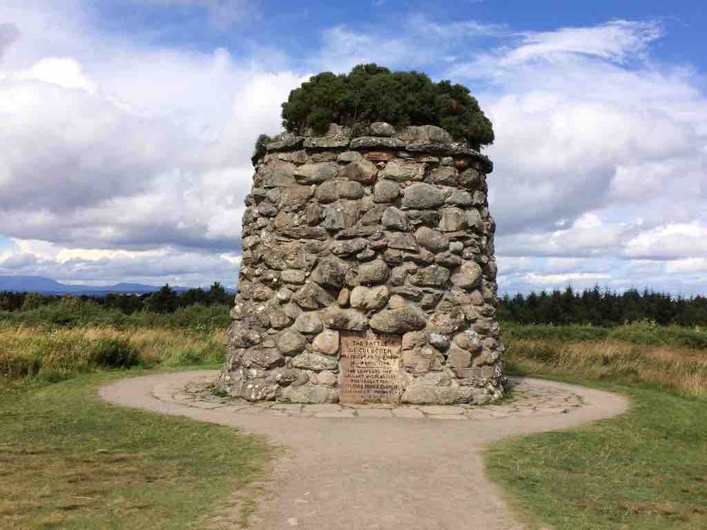 A historic stone monument stands prominently in an open field, surrounded by greenery. The structure features a rounded shape with a leafy top, while an engraved plaque on the front provides information about its significance. Soft clouds drift in a blue sky above.