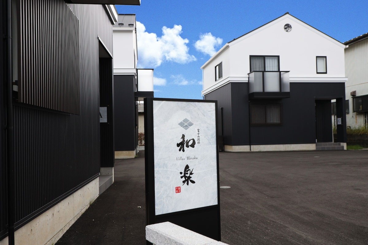 The entrance to the villa is marked by a sign displaying "和楽". Two modern two-story buildings with contrasting dark and white facades are visible in the background. The parking area is spacious and includes a clear blue sky overhead.