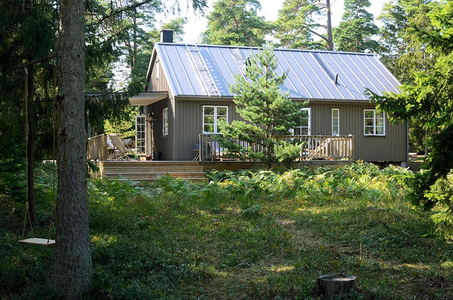 The exterior of the recently renovated beachhouse is surrounded by lush greenery. A spacious wooden deck wraps around the house, providing seating and a peaceful atmosphere. Large windows allow natural light to illuminate the interior, while solar panels are visible on the roof.