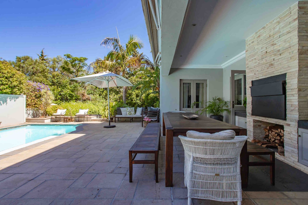 An outdoor dining area features a large wooden table surrounded by wicker chairs. A shaded umbrella stands nearby, and a built-in braai/BBQ is positioned against a textured stone wall. The swimming pool is visible, with loungers arranged around it. Lush greenery adds a natural backdrop.