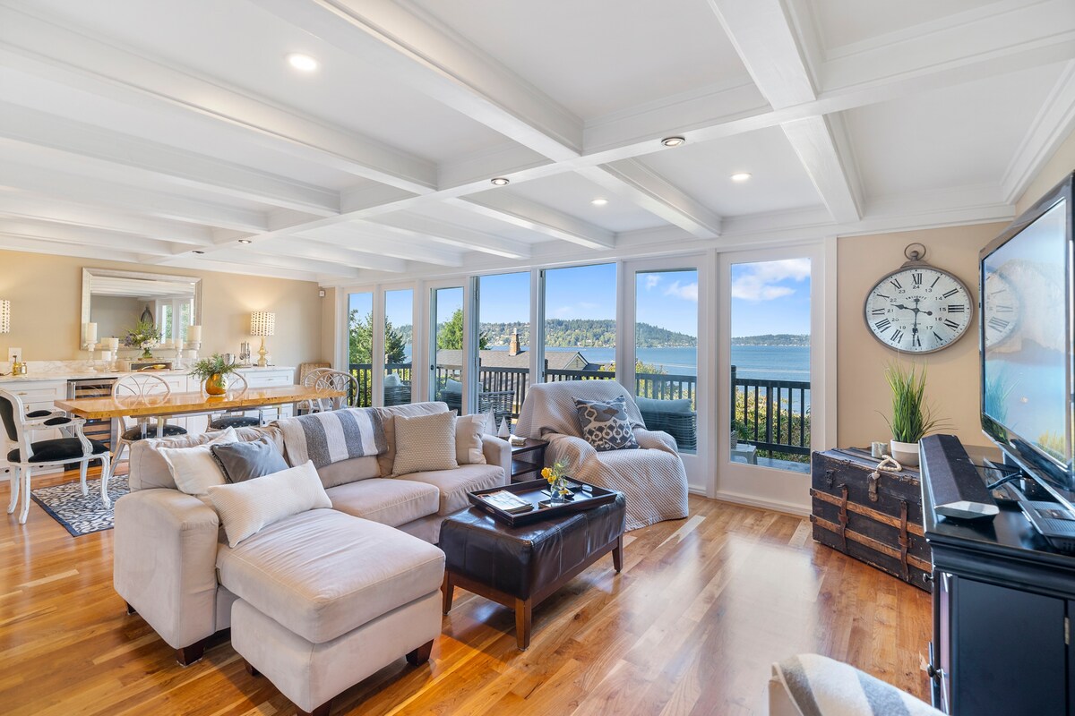 The living area features large windows showcasing expansive views of Lake Washington. Soft furnishings create a comfortable seating arrangement centered around a coffee table. A dining area with a wooden table is visible in the background, while a large clock and TV add functional accents to the space.