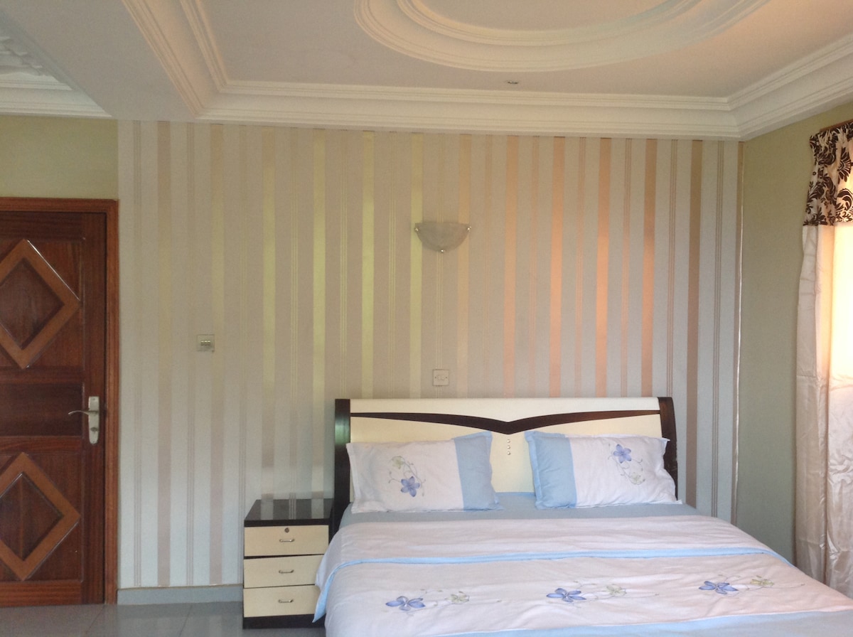 A well-appointed bedroom features a spacious bed with a light blue bedspread and matching pillows. Subtle striped wallpaper complements the decor, while a bedside table with drawers is positioned next to the bed. Natural light enters through a window adorned with decorative curtains.