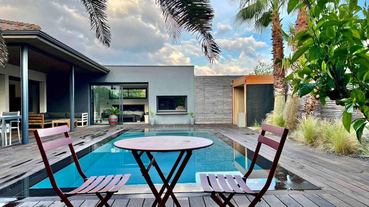 A wooden deck surrounds a modern pool featuring a clear blue surface. Two red folding chairs flank a small round table, inviting relaxation. Lush greenery, including palm trees, provides a tropical feel, while a contemporary structure is visible in the background.