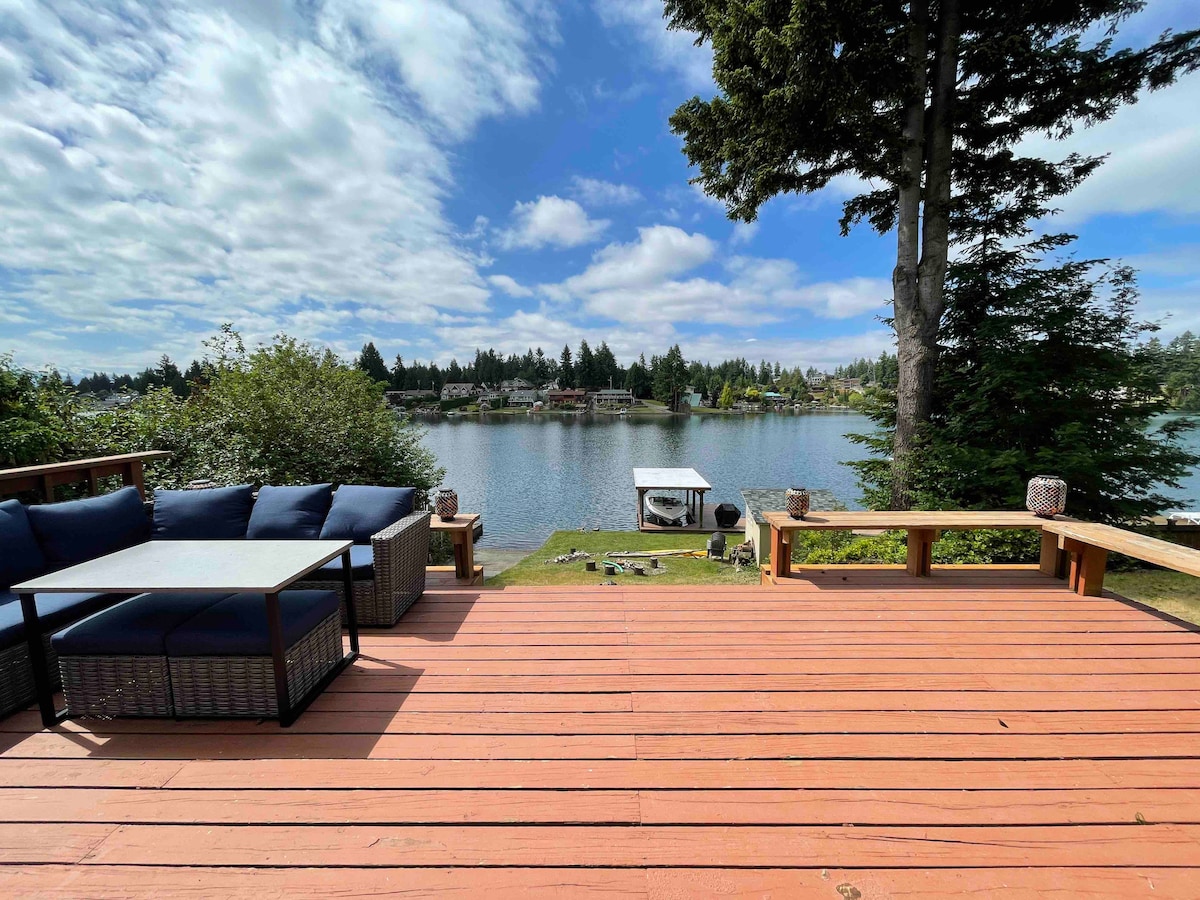 A spacious wooden deck overlooks a serene lake, surrounded by lush greenery. Comfortable outdoor seating is arranged around a coffee table. The landscape reflects the calm water, with a clear blue sky and scattered clouds enhancing the tranquil setting.