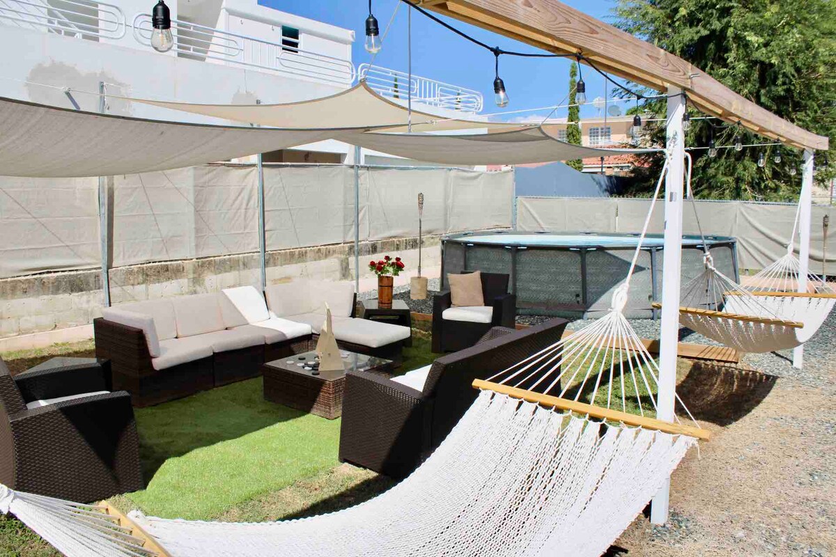 An outdoor seating area is depicted with a brown wicker sectional sofa and a coffee table, shaded by fabric canopies. Two hammocks are positioned nearby, and a circular above-ground pool is visible in the background, surrounded by gravel and greenery.
