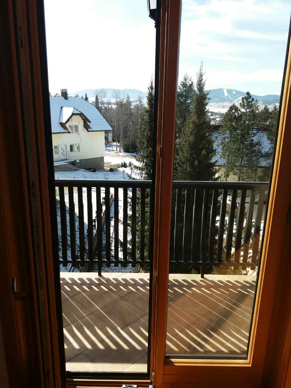 A balcony view framed by wooden doors reveals a snowy landscape and distant mountains. Sunlight casts shadows on the wooden deck. Trees and a neighboring house can be seen, creating a serene outdoor setting.