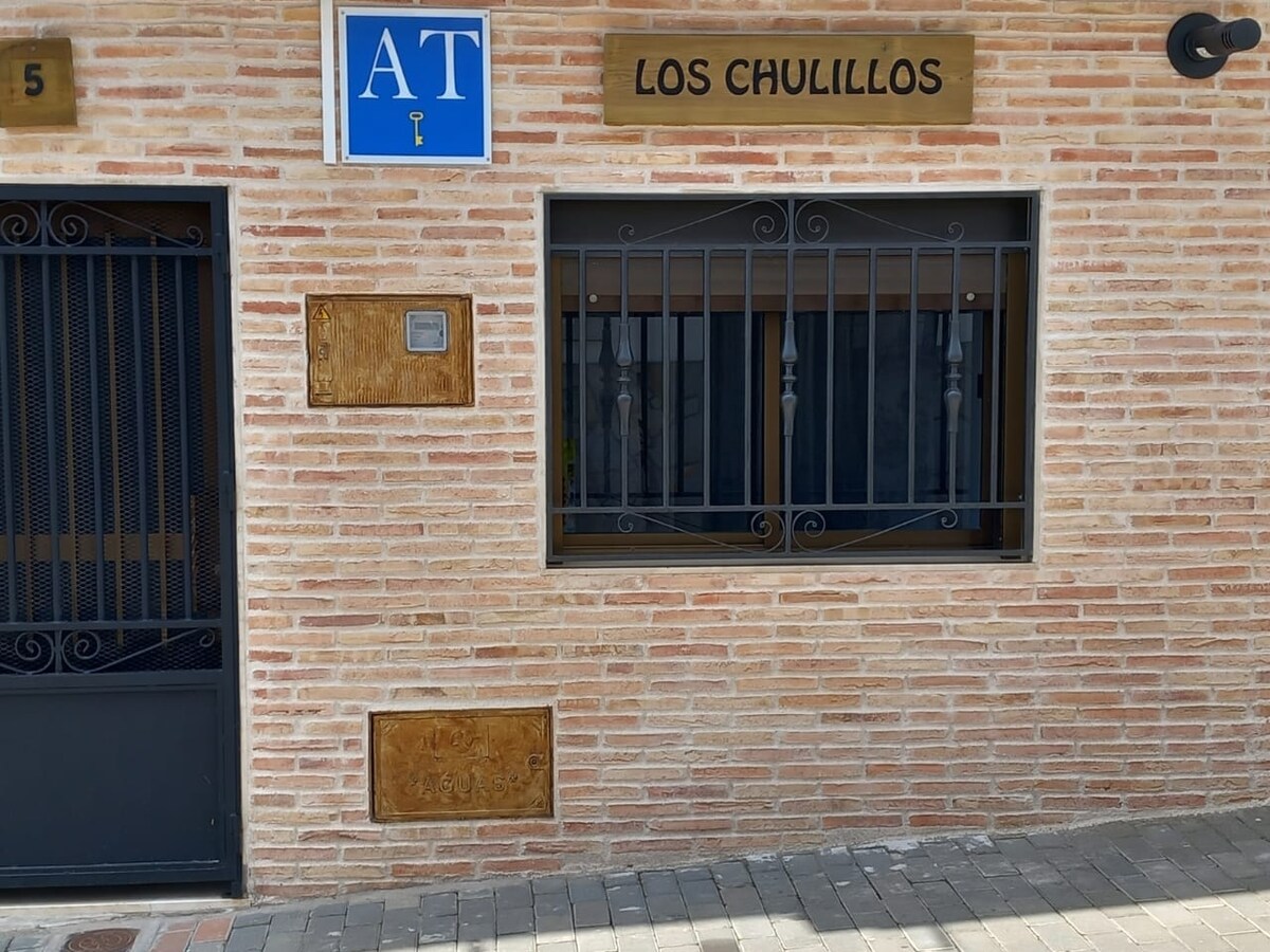 The exterior features a warm brick facade with a decorative sign displaying 'LOS CHULILLOS'. A grid-style window can be seen in the center, enclosed with metal bars. The entry is marked by a simple door, providing access to the accommodation.