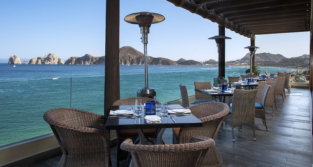 An outdoor dining area is showcased with tables set for meals, overlooking the ocean and distant rock formations. Woven chairs surround the tables, providing a comfortable setting. Heaters are positioned to offer warmth, while the expanse of blue water and clear sky create a serene backdrop.