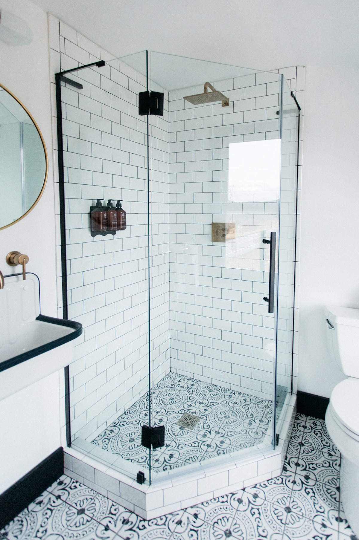 A modern bathroom features a seamless glass shower enclosure with white subway tile and a detailed patterned floor. Natural light filters in, highlighting a round mirror above a sleek black sink, complemented by stylish fixtures and neatly arranged toiletries.