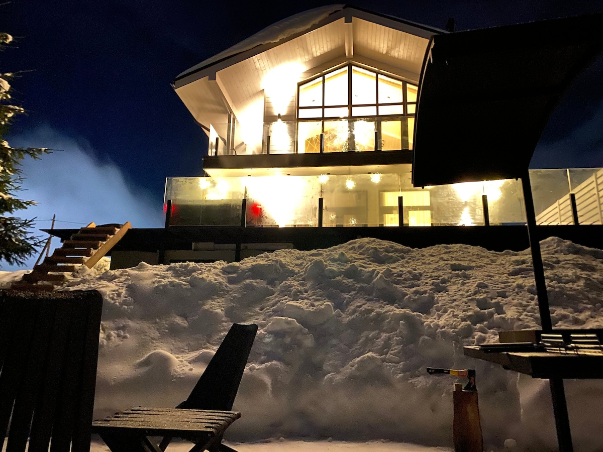 The chalet, illuminated at night, features large windows that reveal a warm interior. A wooden deck sits in front, flanked by snowbanks. A seating area is visible, providing a serene setting against the snowy backdrop.