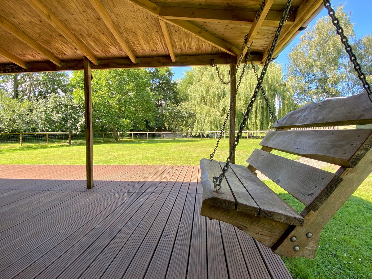 A wooden swing hangs from a covered deck, surrounded by an expansive green lawn. Sunlight filters through the trees, enhancing the serene atmosphere of the garden area. The porch features a smooth, wooden floor that complements the outdoor space.