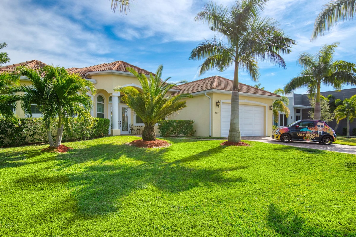 The exterior of a spacious home is depicted, featuring a tile roof and large arched windows. Lush green grass and palm trees surround the entrance, while a driveway leads to a garage. Clear skies with scattered clouds provide a bright and inviting atmosphere.