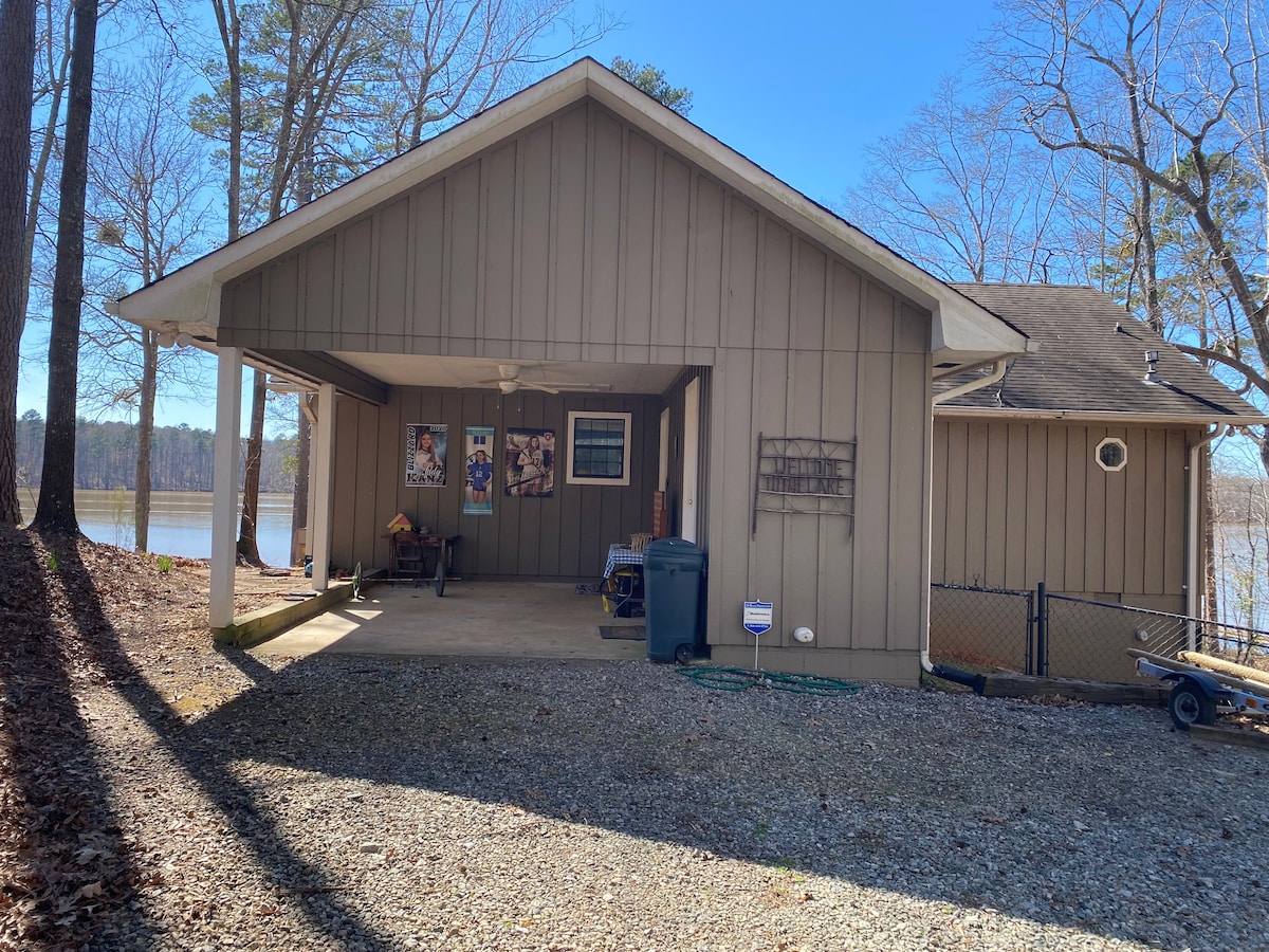 A lakeside bungalow is displayed with a covered porch, showcasing a gravel driveway. The structure features wooden paneling and large windows that overlook the water. Surrounding trees provide shade and add a natural feel to the setting.