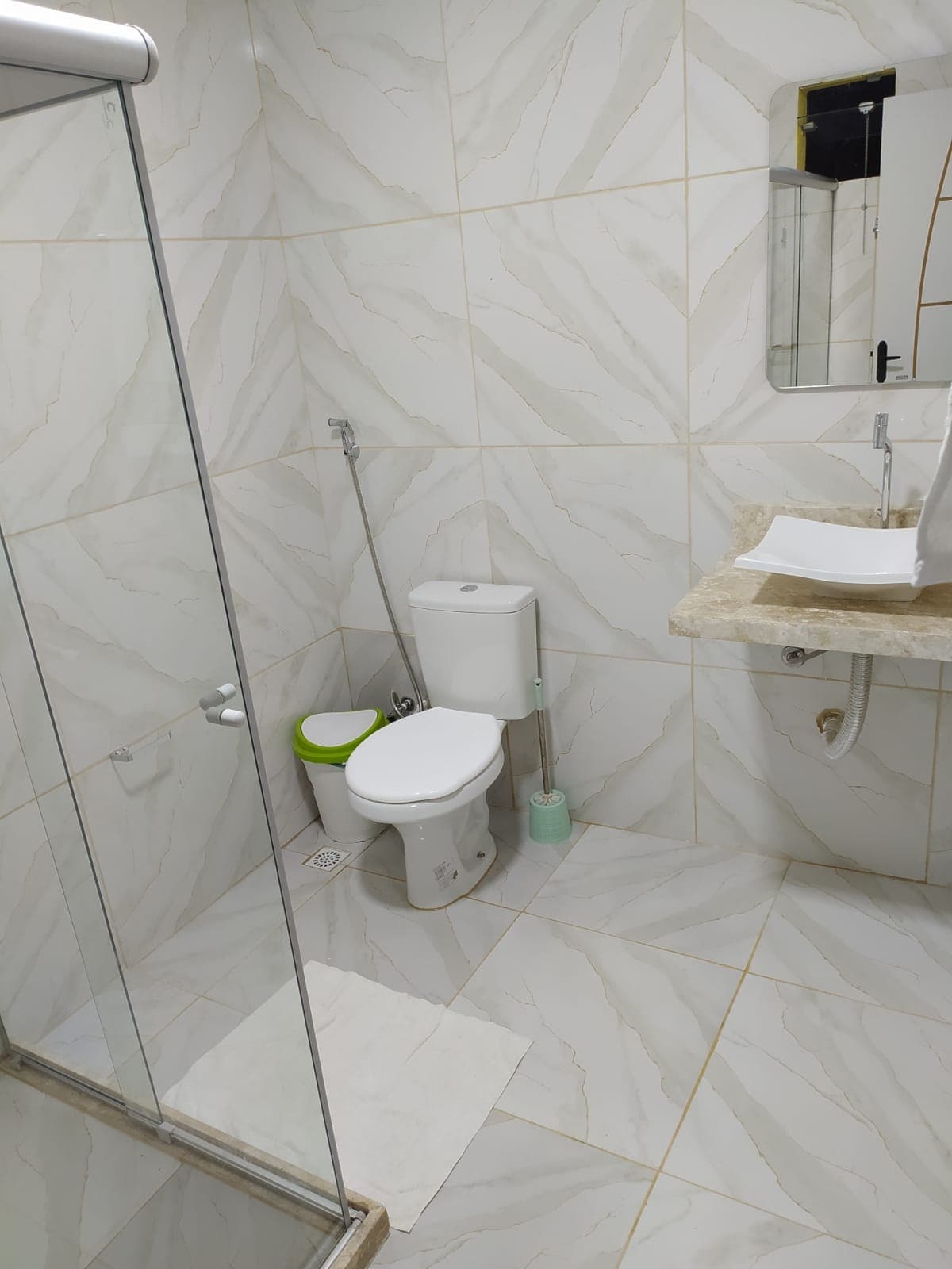 A modern bathroom is presented with light-colored, textured tiles. A glass shower enclosure stands beside a compact toilet and a small green waste bin. A stone countertop holds a white sink, while a mirror reflects the bright space.