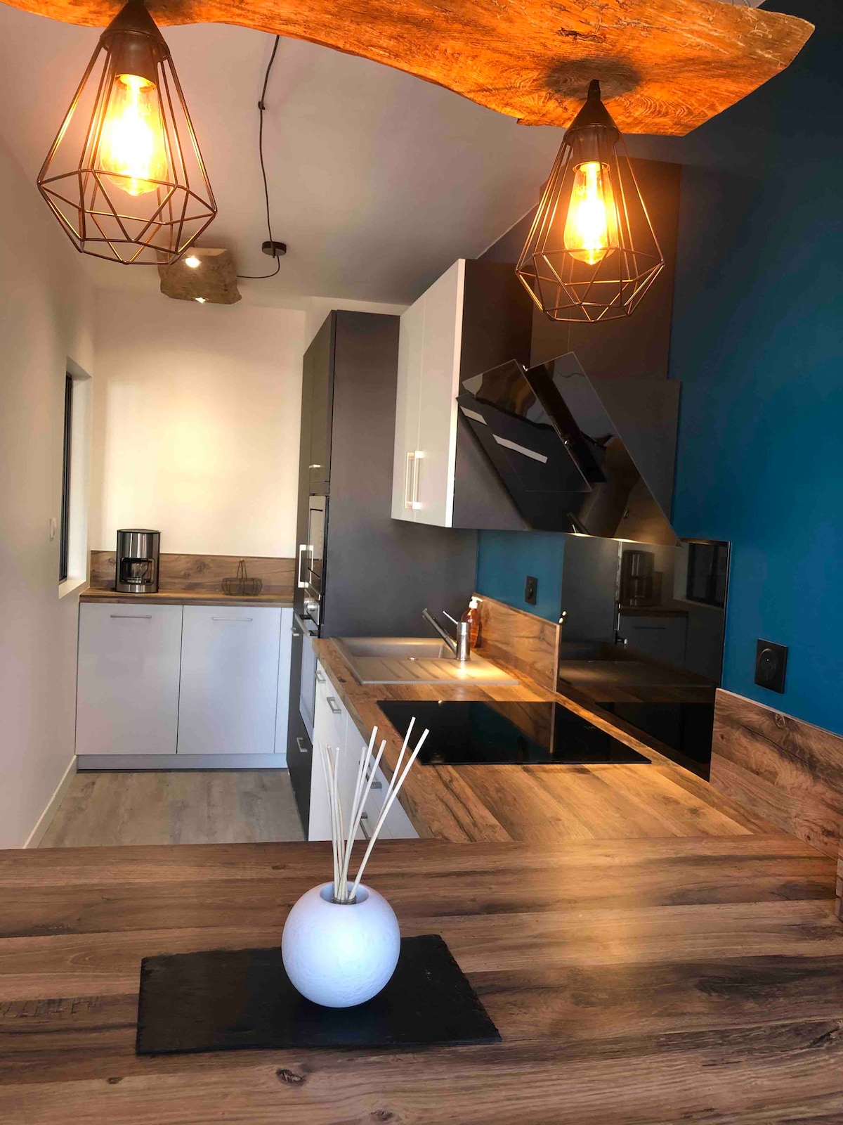 A modern kitchen is presented, featuring a combination of sleek cabinetry and wooden countertops. Two geometric pendant lights illuminate the space, highlighting the dark and light contrasts. A white vase with decorative sticks is visible on the counter, adding a touch of simplicity.