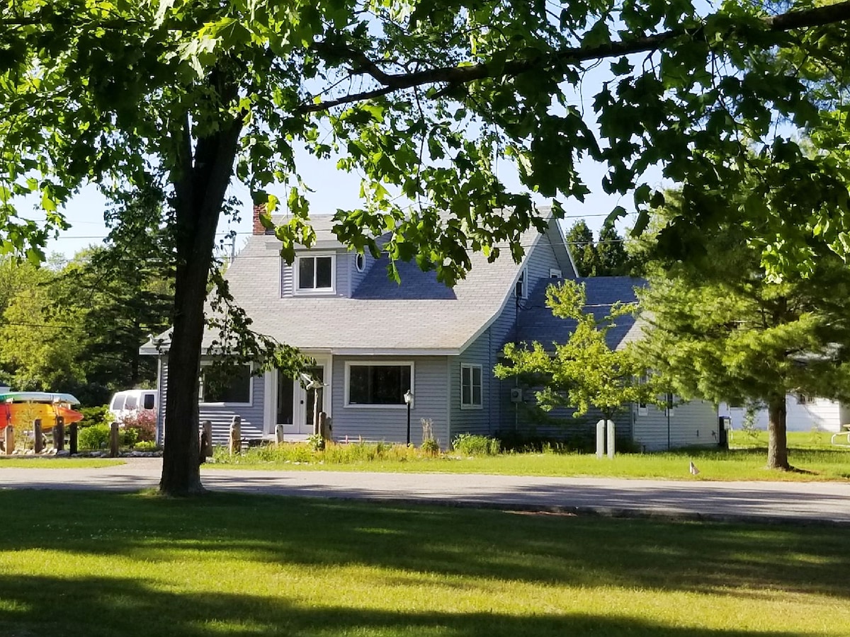 A spacious home is framed by trees, with light gray siding and a contrasting roof. Large windows invite natural light, and a well-maintained lawn stretches in front. The driveway is visible, leading towards the entrance of the house, enhancing the welcoming feel of the property.