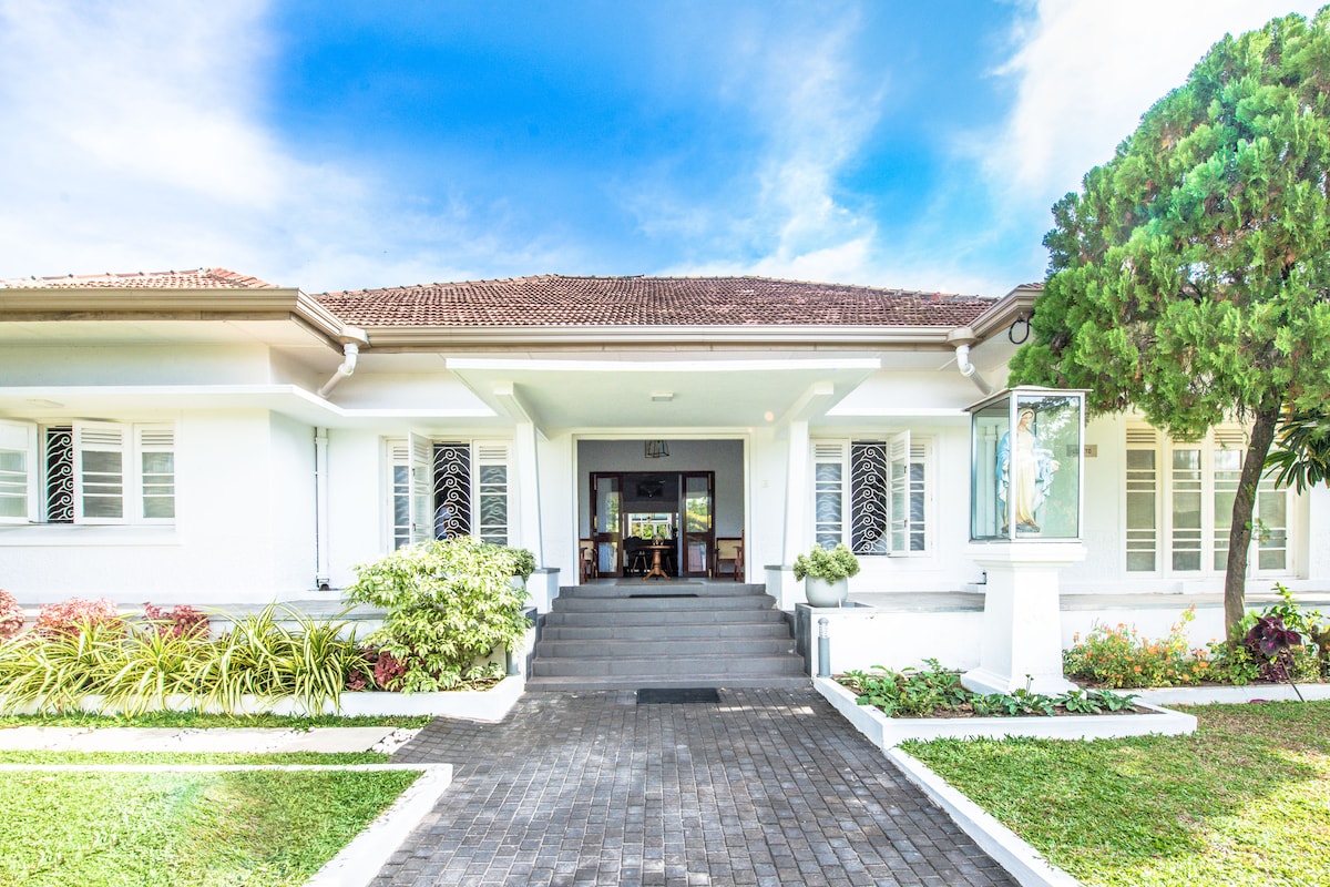The villa's welcoming entrance features a wide staircase leading to the front door, framed by large windows. Lush greenery and colorful landscaping adorn the sides, creating a fresh and inviting atmosphere. A clear blue sky complements the property, enhancing its outdoor appeal.