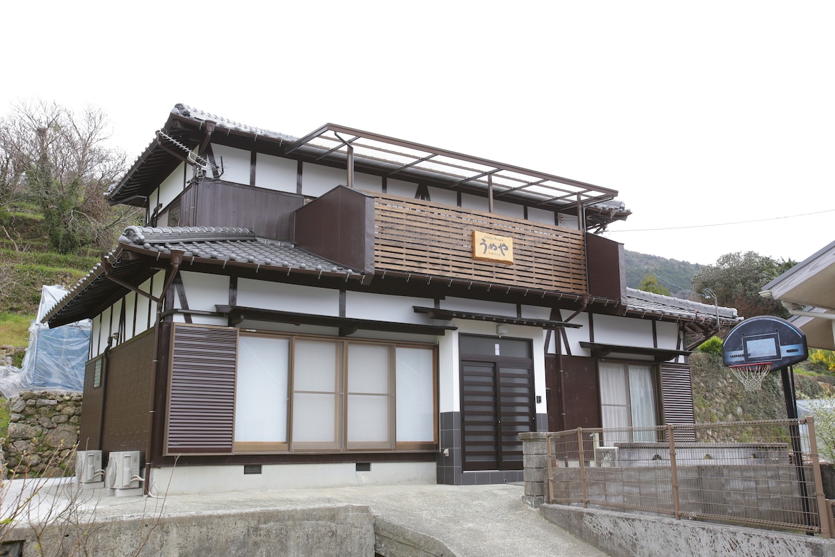 The exterior of a traditional two-story guest house is shown, featuring wooden accents and a sloped roof. Large windows fill the facade, allowing natural light to enter. A unique sign adorns the front, alongside a basketball hoop. The surrounding area is green and hilly.