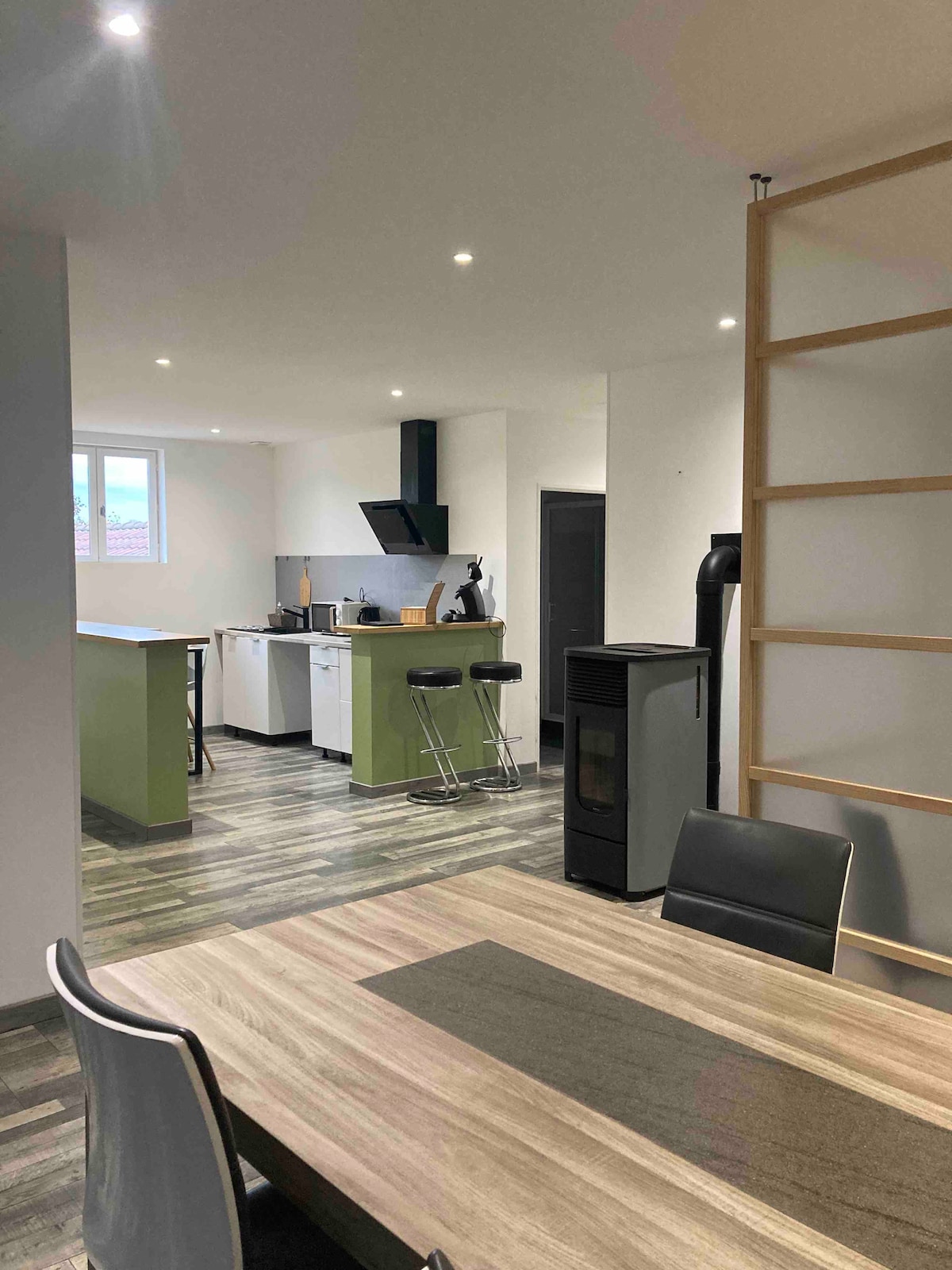 A modern kitchen is visible, featuring sleek cabinetry, a gas stove, and a stylish black extractor hood. A dining table with four chairs occupies the foreground, while a small breakfast bar with two stools is positioned nearby. Large windows provide ample natural light.